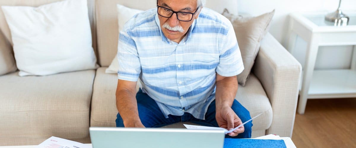 Focused greyhaired elderly man sit on couch reading bank notifications calculating domestic expenditures concentrated modern mature male consider financial paperwork pay bills on laptop online